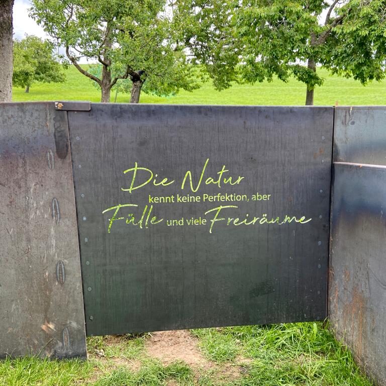 Ein speziell vom Schlosser gebautes Zauntor mit der Aufschrift "Die Natur kennt keine Perfektion, aber Fülle und viel Freiraum".