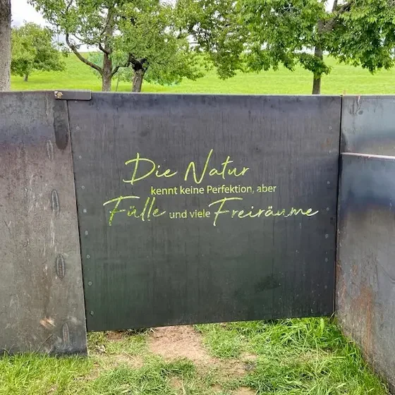 Ein speziell vom Schlosser gebautes Zauntor mit der Aufschrift "Die Natur kennt keine Perfektion, aber Fülle und viel Freiraum".